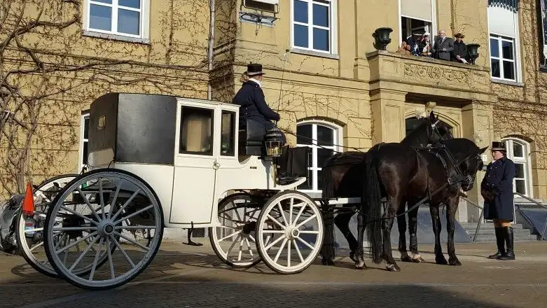 Witte glaslandauer met koetsier en palfrenier op een historisch plein